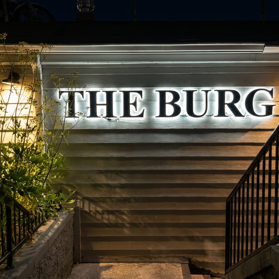 Illuminated The Burg restaurant sign on a wood-paneled exterior, framed by stair railings and greenery at night.