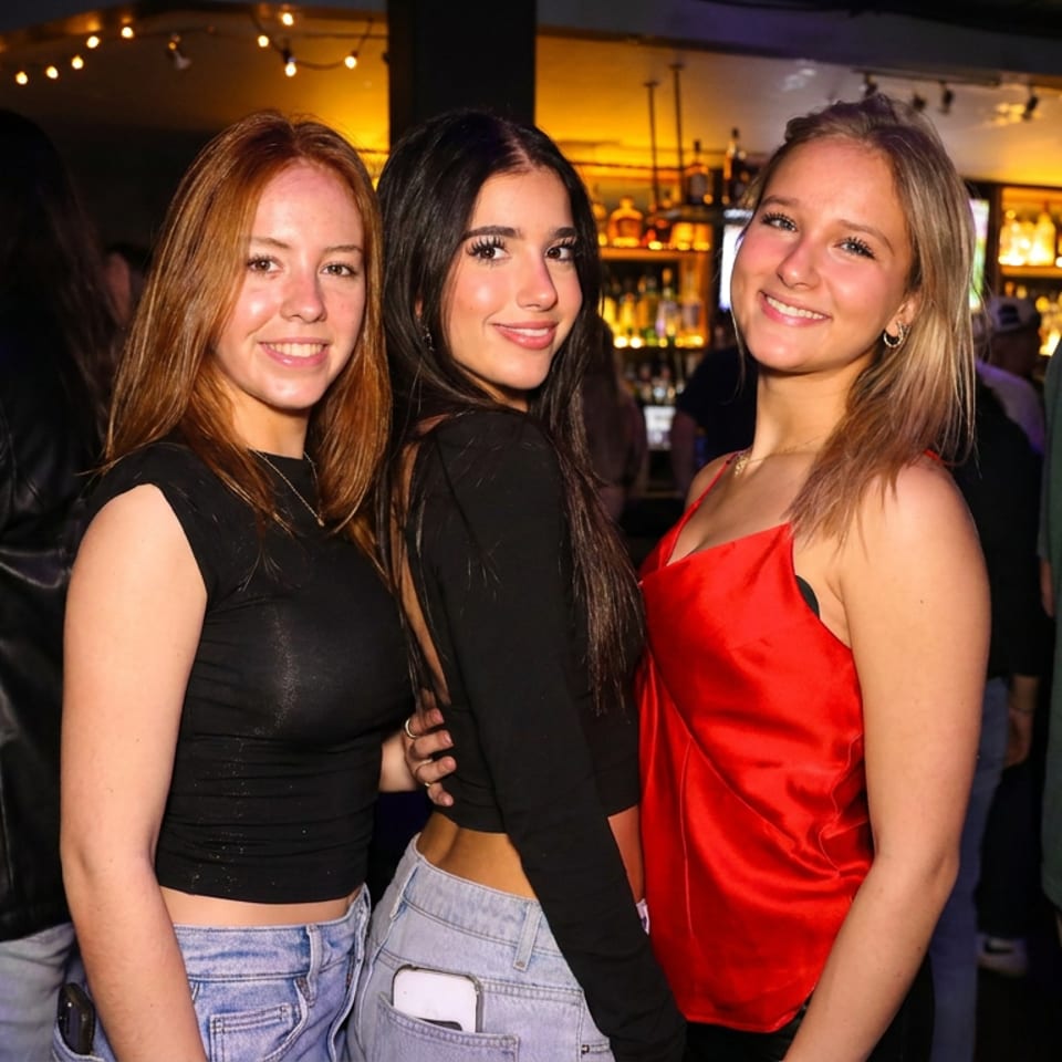 Three women smile together at a lively restaurant bar illuminated by warm string lights, surrounded by other patrons.
