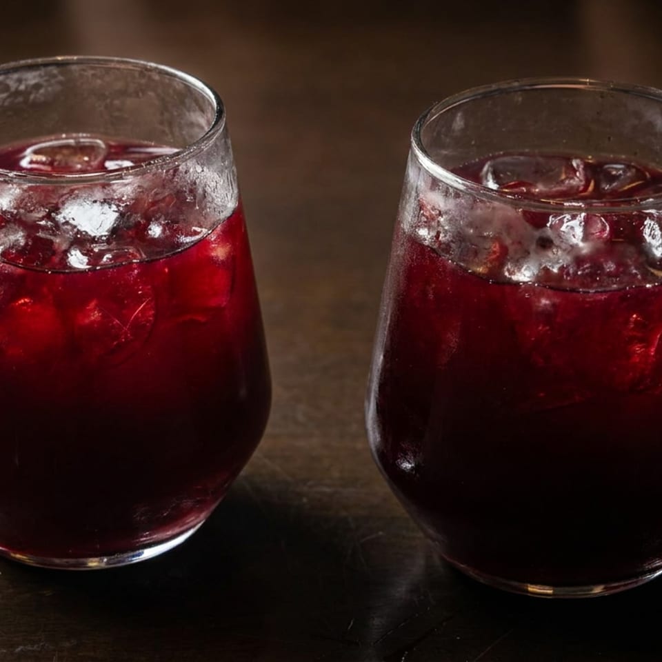 Two glasses of dark red iced drinks sit on a rustic wooden table.