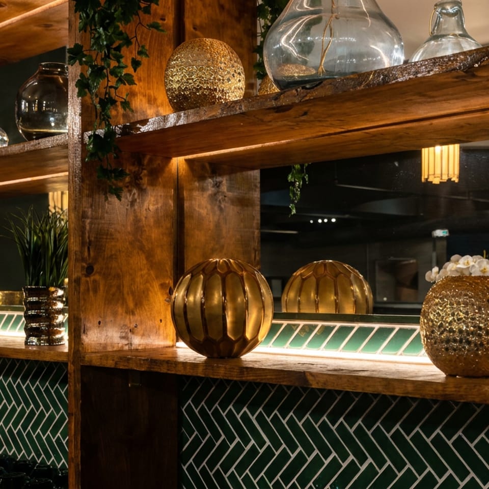 Warmly lit restaurant shelving displays glass vessels, decorative spheres, greenery, and dark green herringbone tile.